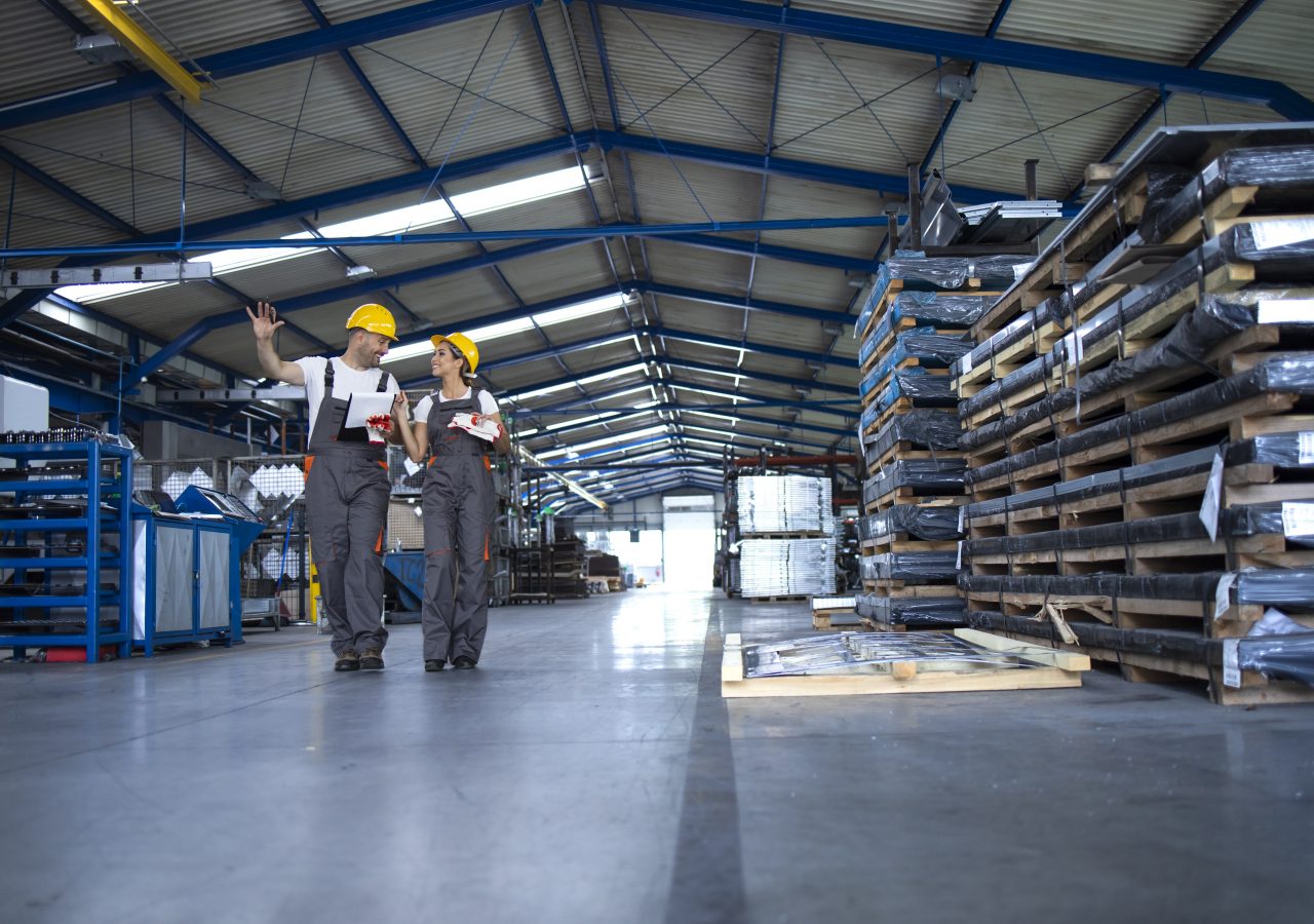 Factory workers in work wear and yellow helmets walking through industrial production hall and sharing ideas about organization.